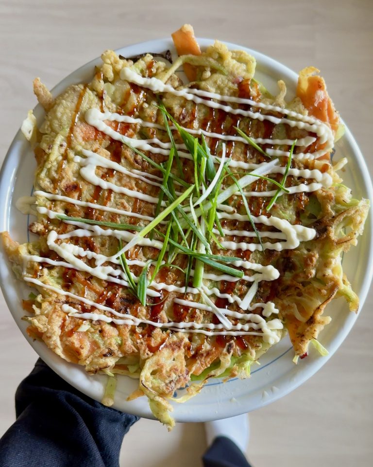 En gyldenstegt okonomiyaki toppet med teriyakisauce, mayonnaise og friske forårsløg på en mørk tallerken.